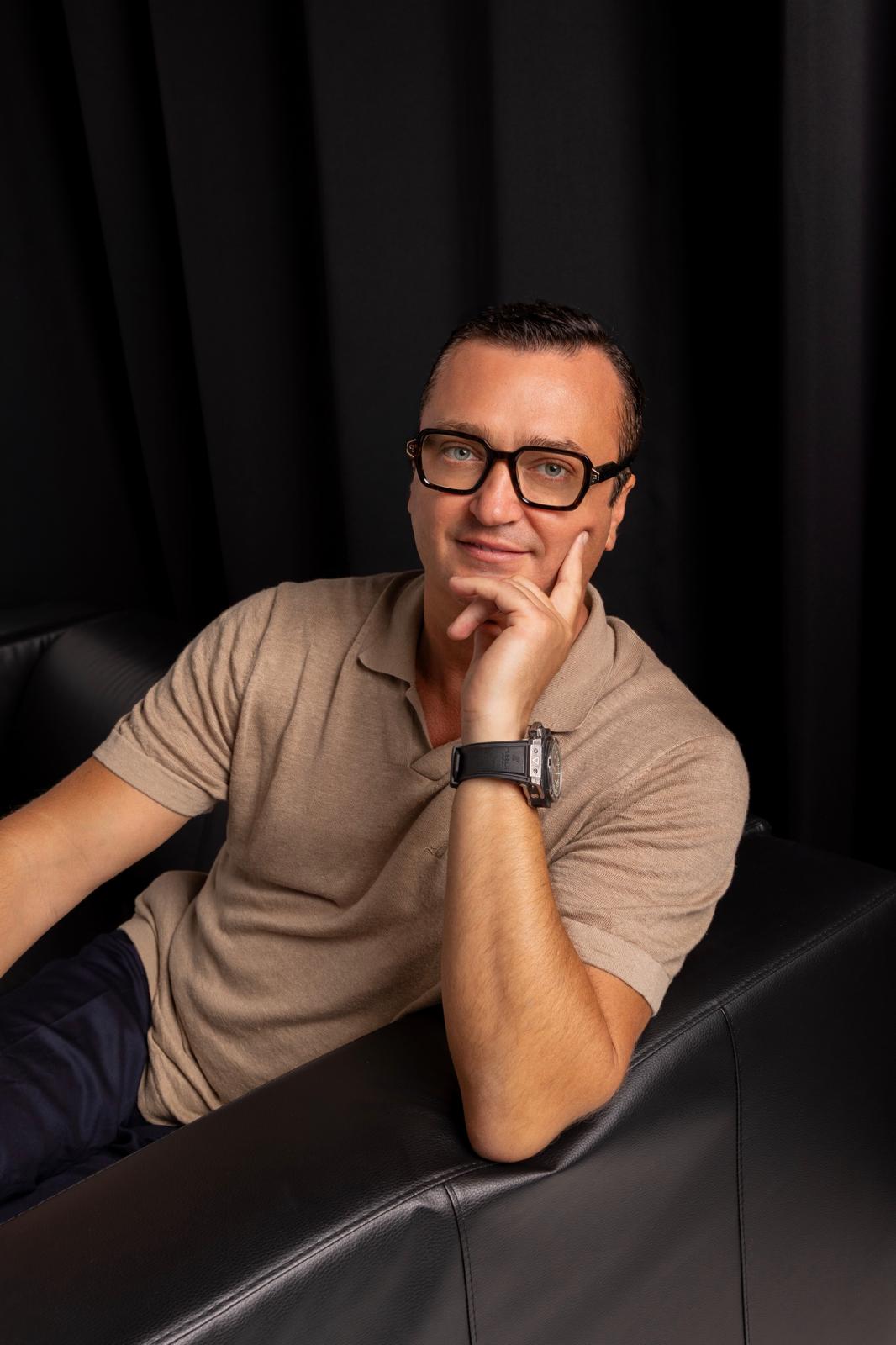 Konstantin Silver in a tan knit polo and glasses, chin resting on his hand, against a black background
