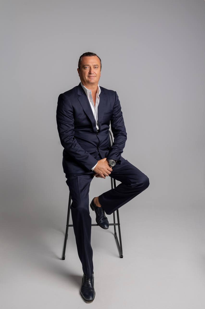 Konstantin Silver in a navy suit seated on a stool against a light grey background