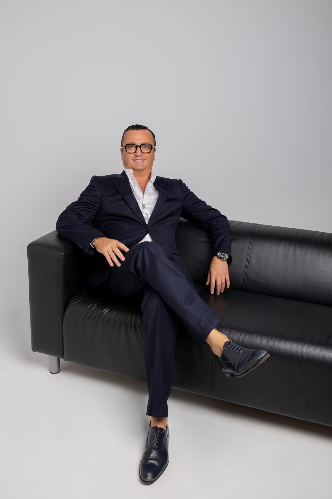 Konstantin Silver in a navy suit seated on a black leather sofa against a light grey background